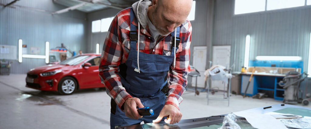 Der männliche Meister in der Werkstatt arbeitet mit der Windschutzscheibe, er benutzt ein Spezialwerkzeug