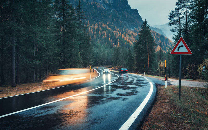 Verschwommene Autos in Bewegung auf der Straße im Herbstwald bei Regen. Perfekte asphaltierte Bergstraße an bewölktem Regentag. Fahrbahn, Pinien in den italienischen Alpen. Transport. Autobahn im nebligen Wald.